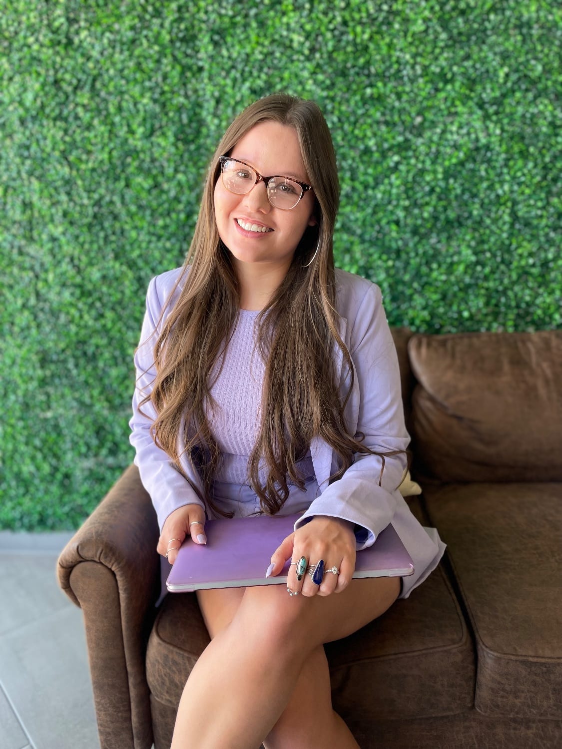 Alissia smiling in a lavender blazer, seated on a brown leather couch in front of a green leaf wall with a laptop on her lap